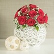 A carefully styled floral composition evokes the atmosphere of a romantic wedding dinner laid out in a converted Clerkenwell warehouse or chic townhouse. Dominating the scene is a tightly formed dome of deep red roses, around a dozen to fifteen blooms, each rose in perfect full flower with richly layered, velvety petals curling inward like delicate silk. They press closely together, creating a sense of abundance and intimacy, as though made for a sweetheart table or a special anniversary. Threaded generously through the roses are sprays of white baby's breath, each cloud of tiny blossoms bringing an airy, almost ethereal softness that tempers the bold red and mirrors lace detailing on a bridal gown. Sparse yet intentional touches of fresh green leaves peek out at the edges, enhancing the natural, garden-fresh feel. The bouquet is nestled in a round white vase with a distinctive open-weave design, as if white wicker or slim rattan has been artfully interlaced into a sculptural nest. Light flows through the gaps, casting faint shadows and adding depth, aligning with the creative, design-forward spirit of Clerkenwell's studios and galleries. To the left, two woven decorative orbs in soft cream and off-white tones rest on the same smooth white surface: one larger, more dominant sphere and a smaller one tucked just behind it, both echoing the airy latticework of the vase. The background is a softly blurred, neutral wall with a gentle fabric-like texture that doesn't compete for attention. Together, these elements create a calm yet passionate mood, perfect for illustrating wedding table flowers, romantic home décor, or local same-day rose arrangements in Clerkenwell.