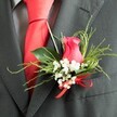 The photograph focuses closely on a single men's wedding boutonniere, fastened neatly to the lapel of a dark charcoal grey suit jacket that appears finely tailored in a smooth wool blend. At its centre sits a rich deep pink rosebud, tipping toward fuchsia, partially opened so the petals curl inward in a soft spiral, velvety and luminous against the darker fabric. Surrounding the rose is a ring of delicate white baby's breath-tiny blooms clustered like a gentle halo, adding an airy brightness that softens the overall look. Lush green foliage supports the design, with broad, glossy leaves at the base and slender, feathery sprigs that stretch outward, giving the arrangement texture and a sense of movement, as if echoing the greenery found in Clerkenwell's pocket parks and hidden courtyards. A red satin ribbon is tied in a small, impeccable bow at the base of the boutonniere, its vivid hue perfectly mirroring the smooth, unpatterned red silk tie lying beneath the lapel. A glimpse of a subtly patterned grey dress shirt completes the layered, coordinated outfit. The lighting is gentle, emphasising the contrast between the rose's vibrant colour, the crisp whiteness of the gypsophila, and the deep tone of the suit, so each detail stands out clearly without harshness. This buttonhole feels ideal for a groom, usher, or special guest arriving at a ceremony near Farringdon or Clerkenwell Green, capturing that mix of tradition and contemporary style, and showing the kind of considered, personal floristry that turns a smart suit into a truly wedding-ready ensemble.