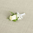 Resting on a softly textured, neutral linen background, this wedding boutonnière captures the kind of understated sophistication that suits a Clerkenwell ceremony perfectly. At its centre, a flawless ivory rose is captured in that tender moment between bud and full bloom, its petals thick, smooth, and layered like satin, creating a gentle spiral of cream against the pale backdrop. Delicate clusters of white baby's breath surround the rose, each tiny gypsophila flower forming a lacy halo that adds lightness and romance, like a drift of cloud over nearby Spa Fields. Fine, curving blades of ornamental grass frame the composition, their vivid green arcs bringing movement and a natural, almost meadow-like feel to this classic palette. The stem is meticulously wrapped in a narrow band of white satin ribbon, wound tight and secure, with a neat, intentional finish that reveals an experienced florist's hand. Light falls softly across the piece, catching on the glossy ribbon and the subtle sheen of the rose petals, while the linen beneath adds a gentle contrast with its understated weave. You can almost sense the cool freshness of the bloom, the crispness of the foliage, and the faint, clean fragrance that would travel just enough to be noticed in a registry office or stylish warehouse venue close to Clerkenwell Green. Perfect for a groom, best man, or cherished guest, this boutonnière feels like a heartfelt detail, crafted with care for a day when every small element matters.