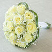 As a florist who has delivered to St John's Square and Exmouth Market many times, I describe this bridal bouquet as a study in calm, classic beauty shaped for a Clerkenwell celebration. About fifteen to twenty creamy yellow roses form the generous, rounded centre-each bloom showing the familiar spiral of petals that opens into an ivory heart with soft, luminous highlights. Wisps of tiny white Gypsophila nestle between the roses, creating a light, feathery veil that contrasts gently with the fuller rose heads and lends an ethereal softness to the whole. Around the edge, long, vibrant green blades-likely lily grass or bear grass-arc with a poised, sculptural curve, almost cradling the roses into a natural frame. At the base, the stems remain visible and are tightly bound with pristine white satin ribbon, knotted with the same care we apply to every bouquet at Clerkenwell Flowers; the wrapped handle reads cool and secure in the hand. The arrangement is photographed on a pale taupe linen surface which complements the cream tones and allows the subtle variations in petal colour to show through under diffuse, soft lighting. There's a subtle, clean fragrance of fresh rose and linen suggested by the image, a quiet confidence that suits weddings, vow renewals or elegant anniversary gestures around Farringdon or Leather Lane. This image highlights expert hand-tying, balanced composition and a timeless palette-an understated, romantic choice for anyone seeking seasonal freshness and refined simplicity in central London.