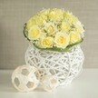 A lush, rounded bouquet of soft yellow roses rests in a contemporary white woven vase, creating a serene yet sophisticated focal point that would feel at home on a wedding table in one of Clerkenwell's converted warehouse venues. Each rose is fully open, the petals forming plush, layered whorls in shades of buttery yellow and cream, pressed closely together to create a smooth, dome-like surface. Scattered throughout the roses, delicate stems of white baby's breath emerge, their tiny blossoms adding a gentle froth, like a fine mist settled over the blooms. Just beneath the flowers, a narrow band of vivid green foliage, possibly flat woven leaves, encircles the base, giving the design a neat, tailored finish and emphasising the flowers' soft tones. The arrangement rises from a striking white vase made of interwoven strands, its openwork, rattan-like structure forming a rounded shape that subtly pinches at the middle, allowing light to dance through the gaps. On the gleaming white tabletop beside it are two decorative spherical accents, created from slender, pale strips of natural material arranged in an airy lattice that echoes the vase's texture. Their placement, one slightly in front of the other, adds depth and a relaxed, modern rhythm. The background is a softly blurred field of beige or light taupe, reminiscent of a calm interior wall in a Clerkenwell apartment, ensuring all focus remains on the floral artistry. The effect is one of quiet celebration and refined taste, ideal for contemporary wedding centrepieces, intimate dinner parties, or stylish same-day delivery across Clerkenwell and nearby Farringdon.