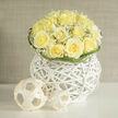 This arrangement reads like a symphony in ivory and pale butter-creamy yellow roses clustered into a perfect dome, their ruffled centers unfurling in layered softness. Wisps of baby's breath drift between the petals, lending a delicate, dancing lightness that softens the bouquet's overall silhouette. The flowers are cradled within a striking white wicker orb: a modern vessel whose crisp lines and open weave both contain and reveal the blooms, creating tactile intrigue and a contemporary flourish. The composition is set in soft, natural daylight that catches the roses' subtle tonal variations, from buttery heart to palest edge, while the gypsophila catches tiny points of reflected light like distant stars. Touch and scent converge-the velvety petals, the slight crunch of woven wicker, and a fragrance that nods to fresh-cut lawns and sun-washed linen with a faint earthy undertone. Curved blades of greenery loop at the rim, their controlled gestures framing the arrangement and guiding the eye inward. Simple woven spheres placed beside the basket introduce a sculptural, playful counterpoint that emphasizes purity of form. This bouquet suits moments of hope and congratulations-a thoughtful gift for friends returning to Clerkenwell after a hospital stay, a centrepiece for a quiet anniversary dinner near Exmouth Market, or a soft, uplifting welcome to a studio on Clerkenwell Green. As arranged by a florist acquainted with the district's calm, considered taste, it balances contemporary design with traditional softness and invites a measured, joyful response.