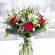 A luxurious red and white bouquet sits proudly in a clear cylindrical glass vase, its neatly cut green stems visible through the fresh water, giving an immediate sense of care and vitality. At the heart of the arrangement, velvety deep red roses open slowly, their layered petals curling outward in soft spirals that speak of timeless romance. A large, deeply ruffled crimson carnation nestles near the front, its intricate, feathery texture adding drama and depth to the design. Around these passionate reds, clusters of pure white blooms create a calm, luminous contrast: delicate white alstroemeria with slim, speckled petals, creamy chrysanthemum-style flowers, and several closed white lily buds that promise to unfurl in the days ahead. Sprays of tiny white gypsophila float like a fine mist between the larger flowers, giving the whole bouquet a light, cloud-like softness. Lush greenery wraps and frames the composition, from broad, glossy dark green leaves carefully curved around the base like an elegant collar, to eucalyptus-style foliage and pointed stems that reach upward and outward. The bouquet rests on a pristine white surface, with gentle natural light-much like the daylight drifting through Clerkenwell studio windows-washing over every petal and leaf. The background is softly blurred, keeping all attention on the flowers while still suggesting a bright, welcoming interior. The mood is polished yet heartfelt, ideal as a centrepiece for a celebration dinner near Exmouth Market, an anniversary surprise in a Clerkenwell townhouse, or a thoughtful gift to brighten an office near Farringdon, carrying feelings of admiration, love, and warm congratulations.