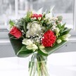 A contemporary glass-vase bouquet glows in soft daylight, as if catching the calm of a Clerkenwell morning. At the heart of the arrangement, a single coral-red rose and a plush crimson carnation provide a rich, velvety focal point, their layered petals full and dimensional. Around them, clusters of small white chrysanthemums, daisy-like blooms, and tiny button flowers create a halo of brightness, each petal crisp and clean against the deeper tones. Creamy white lilies, some just beginning to open and others still in smooth, tapered buds, rise gracefully through the design, hinting at fragrance yet to unfold. The flowers are cradled by wide, glossy emerald leaves, carefully folded and curved so they arc around the base, giving the bouquet a sculpted, almost architectural silhouette. Clear water shimmers through the transparent glass vase, catching reflections from the window and adding a sense of freshness and movement. The overall mood is one of gentle celebration and airy elegance, like something you might spot on a windowsill overlooking Exmouth Market or a quiet side street near St John's Square. You can almost imagine the subtle, sweet scent mingling with a faint green freshness, the textures ranging from silky rose petals to the soft fluff of chrysanthemums. This is the kind of floral arrangement that would sit beautifully on a dining table for a birthday, an anniversary, or simply to brighten a Clerkenwell flat after a long day, radiating thoughtful design and a quietly joyful energy.