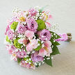 Laid gently on a pale grey, linen-like cloth, this lush hand-tied bouquet feels like a still moment before a wedding begins somewhere in Clerkenwell's cobbled backstreets. The flowers form a dense, rounded dome of colour where lavender roses take centre stage, their petals opening in generous spirals that create a soft, plush texture. Between these, pink alstroemeria unfurl with elegant, slender petals brushed with blush, cream, and flashes of yellow near their throats, each floret carrying fine speckles that mimic delicate brushstrokes. Nestled throughout the arrangement, small, perfectly rounded button chrysanthemums glow in bright chartreuse green, injecting energy and contrast, as if echoing the fresh vibrancy of nearby Exmouth Market on a spring morning. Tiny clusters of white baby's breath are woven between every bloom, light as mist, giving the bouquet a gentle, romantic haze and softening the transitions from one flower to the next. At the base, the stems are carefully aligned and tightly bound with a broad band of pink satin ribbon, its sheen catching the light, while a slim strand of white pearls spirals over the handle, adding a bridal detail reminiscent of a cherished necklace. The natural stems extend just beyond the ribbon wrap, bringing an earthy, organic finish that would feel at home in a ceremony at a converted Clerkenwell warehouse or an intimate dinner along St John Street. The bouquet's mix of pastel roses, lively greens, and airy white accents conveys hope, tenderness, and celebration - an arrangement that suits engagements, registry weddings, or milestone anniversaries delivered across Clerkenwell.