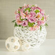 A polished, close-up scene presents a rounded wedding-style arrangement that seems perfectly at home in Clerkenwell's blend of heritage and modern design. The bouquet forms a gentle dome of colour, dominated by lush lavender and blush-pink roses, each bloom in full, velvety glory, their petals unfolding in delicate spirals that catch the light. Threaded among them are elegant stems of pale pink alstroemeria, their elongated petals etched with fine speckles and soft streaks of deeper pink and apricot, like tiny brushstrokes on silk. Lively lime green button chrysanthemums provide a fresh, contemporary contrast to the pastels, their neat, spherical heads giving a playful yet structured feel. Between the larger flowers, airy sprigs of white baby's breath drift like light mist, softening edges and lending a dreamy, romantic quality. Dark green foliage tucked low in the arrangement offers depth and frames the pastel tones. All of this is cradled in a white spherical vase woven from wicker or rattan, with an open, lattice-like construction that makes the base look almost like a sculptural lantern. Two smaller woven orbs, in paler, natural fibres, rest nearby on the same flawless white surface, echoing the woven texture and adding a thoughtful decorative touch. The background is a simple, neutral wall in muted beige or off-white, subtly textured and unobtrusive, much like the understated interiors of Clerkenwell flats and studios. The soft, even lighting suggests natural daylight streaming through a nearby window, creating an atmosphere of calm celebration-ideal for a wedding breakfast table, a registry office bouquet display, or a refined engagement gathering just off Farringdon Lane.