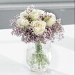 A close-up image shows an elegant white and lavender bouquet resting on a bright white tabletop, the whole arrangement bathed in soft natural light that feels like a calm Clerkenwell morning. At the heart of the design are several creamy white roses, their petals flawlessly smooth and gently unfurling, with a faint blush of pale yellow deep in the centre that suggests they have just opened. Each rose appears full and velvety, the kind of bloom you instinctively want to lean in and breathe in, with a subtle, fresh fragrance that recalls spring walks through nearby Spa Fields or St James's Church Garden. Surrounding the roses is a cloud of lavender-hued statice or limonium, its tiny clustered flowers forming a delicate, almost mist-like halo around the focal blooms. The statice has a papery yet airy texture, giving the bouquet volume without heaviness, and its soft purple tones lend a peaceful contrast to the purity of the white roses. All the stems sit in a clear glass vase with a distinctive crackled, ice-like surface; the textured glass catches and scatters the daylight in fine glints and reflections, adding quiet sparkle without distracting from the flowers. Through the water inside, you can see the straight, fresh green stems, cleanly cut and neatly arranged by an experienced florist, anchoring the composition. The background fades away into a gentle blur of pale grey, keeping every bit of attention on the serene, understated beauty of the bouquet. The overall mood is tranquil and comforting, perfect for a thoughtful sympathy gesture, a gentle get-well arrangement delivered to a Clerkenwell studio, or simply to bring a sense of quiet elegance to a minimalist London home or office.