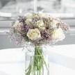 A round, luxuriously full bouquet of creamy white roses and lilac-tinted statice stands quietly radiant at the centre of the image, captured as if it has just been placed on a calm, sunlit table in Clerkenwell. About a dozen white roses form the heart of the design, each bloom perfectly formed, with velvety petals unfolding in soft spirals from warm, creamy centres to gently ivory tips. They sit close together, creating a plush, cloud-like dome that suggests classic romance and reassuring calm. Threaded among the roses, countless tiny blossoms of pale lavender statice weave an airy haze, their papery petals and fine branching stems giving the bouquet a delicate, misty halo. The cool lilac tones play beautifully against the creamy whites, adding depth and a hint of contemporary style without disturbing the overall serenity. All the stems gather neatly into a tall, clear cylindrical glass vase, the fresh green stems visible beneath the water line, some crossing like fine lines drawn in soft pencil. The vase rests on a smooth white surface that bounces the daylight upwards, brightening the underside of the blooms and adding a subtle reflection. In the softly blurred background, the suggestion of a large window lets gentle natural light wash through the scene, much like the light you find in a Clerkenwell studio or apartment on a clear morning. The petals seem to glow from within, while the statice casts faint shadows, creating a feeling of quiet hope and reflective tenderness. This bouquet feels ideal for sending sympathy, marking an anniversary, or offering a peaceful, elegant gesture of support to someone nearby in EC1, whether at home, in a calm office, or in a quiet corner overlooking Spa Fields or Exmouth Market.