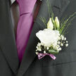 A wedding boutonnière, designed with the quiet precision of an experienced Clerkenwell florist, rests against a dark, well-cut lapel, its details revealed gradually as the eye lingers. At the centre is a flawless white lisianthus, its petals layered like fine fabric, gently ruffled at the edges and opening in a soft spiral that conveys tenderness and calm. Two elegant buds stand just behind the main bloom, their pale green casings still closed, suggesting the sense of new beginnings that suits a ceremony in one of Clerkenwell's historic churches or registry offices. Around them, tufts of delicate baby's breath float like tiny clouds, filling the negative space with lightness and air, each minuscule blossom catching the light as though touched by morning sun filtering through nearby townhouse windows. Slender, variegated foliage weaves through the arrangement, adding flickers of fresh green that break up the white and introduce an organic, slightly wild note, as if gathered from a city garden off Rosebery Avenue. A single deep green leaf curves gracefully at the base, cradling the stems and giving the composition a sense of intention and structure. The finishing touch is a narrow mauve ribbon, tied into a neat, unobtrusive bow that sits snugly against the suit fabric and subtly complements a coordinating tie or pocket square. The boutonnière feels small but deeply considered: you can almost sense the soft brush of petals against the jacket each time the groom moves, and imagine a faint, clean floral scent following him up the aisle. It's a refined, heartfelt accent that captures both the romance of the day and Clerkenwell's modern, creative character.