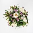 A richly detailed overhead photograph shows a compact, hand-tied bouquet laid on a flawless white background, the clean surface echoing the bright studio light that makes every petal crisp and distinct. At the heart of the design, several creamy white and soft ivory roses open in gentle spirals, some still slightly cupped, others fully unfurled so that their velvety layers form perfect rosettes. Their colour holds just a whisper of warm pale butter at the centres, suggesting freshness and the quiet scent you'd expect from roses gathered that same morning from a trusted supplier. Threaded between these focal blooms are alstroemeria in cool, romantic tones: mauve, lavender, and muted berry-pink petals, some still in smooth, tapered buds, others beginning to open into six-pointed stars gently brushed with green at the tips or streaked with darker markings. Fine clouds of pure white gypsophila weave through the composition, each tiny flower on its wiry stem catching the light and softening any hard edge, so the bouquet feels almost weightless despite its fullness. Beneath and around the flowers, broad, glossy leaves in a rich, deep green fold and curve to cradle the blooms, giving structure and a sense of grounded calm. The arrangement feels like something a Clerkenwell florist might place on a polished table in a converted warehouse flat or a design studio off St John Street: modern, quietly luxurious, and composed with an intuitive eye for balance. Its mood is one of tender celebration and gentle solace, equally suited to a birthday, an understated wedding, or a thoughtful delivery to someone in Clerkenwell who needs a moment of light.