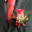 This image shows an elegant wedding boutonniere pinned neatly to the lapel of a dark charcoal suit, the kind you might see at a registry office ceremony near Clerkenwell Green. At its heart, a single deep pink rose opens in gentle spirals, each petal richly coloured and softly curved, giving the bloom a look of calm confidence and romantic depth. The rose is not overly large, but perfectly proportioned, anchoring the design with a classic beauty that feels timeless rather than fussy.

Surrounding the central flower, clusters of white gypsophila-baby's breath-appear like light, airy lace, filling the small spaces with tiny, cloud-like blossoms. These fine flowers lend an ethereal, almost misty quality, softening the strong pink of the rose. Threaded between them, glossy red hypericum berries offer an unexpected twist, their bold, rounded shapes bringing a sense of cheerful energy and a subtle, berry-like sheen when the light catches their surfaces. Wisps of fresh greenery peek out between the berries and gypsophila, their slim, bright green leaves adding a sense of life and movement, as if the piece had just been gathered from a garden corner off St John Street.

All of this is neatly cupped in a single looped leaf, its deep green surface smooth and gently arched, providing structure and a modern line that mirrors the sharp tailoring of the suit. At the base, a narrow red ribbon is wrapped and tied with precision, its colour echoing the berries and adding a final note of celebration. The overall impression is one of poised romance and quiet anticipation, a handcrafted detail that would suit a groom or wedding guest attending an intimate celebration in the heart of Clerkenwell.