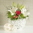 A contemporary yet romantic floral centrepiece is displayed on a pristine white surface, set against a softly textured neutral wall that evokes the calm interiors of Clerkenwell's converted warehouse venues. At the base of the design sits a distinctive white round vase, crafted from interwoven strands that form an open, basket-like lattice. The light passes through this structure, creating subtle shadows and giving the composition a sculptural foundation. The vase overflows with a luxurious mix of wedding-appropriate blooms: stately white lilies rise above the arrangement, some fully open to reveal their broad, elegant petals and warm yellow stamens, others resting in sleek green buds that promise fragrance and beauty throughout the event. Tucked between the lilies is a standout red rose, its velvety petals tightly layered in a classic spiral, offering a strong focal point of deep, romantic colour. Bright green, cushiony pom-pom flowers bring a fresh, modern accent, their spherical shapes contrasting beautifully with the lilies' long lines. Fine clusters of white gypsophila weave through the design, softening the transitions between blooms like a delicate mist, while small clusters of red berries introduce an additional hint of seasonal warmth. A mix of foliage, from feathery, trailing fronds to broader, darker leaves, spills outward, giving the arrangement shape, movement, and a lush, garden-like feel. To the left of the main vase, two decorative spheres, woven from pale natural material, echo the same open pattern, tying the scene together visually. The overall effect is of a thoughtfully curated wedding table arrangement, perfect for receptions and celebrations in and around Clerkenwell, where understated elegance and contemporary design meet.