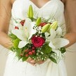 A breathtaking bridal bouquet, handcrafted in Clerkenwell, is elegantly framed against a bride's white, subtly patterned wedding gown, set aglow in soft natural light. The pièce de résistance of this arrangement are the pure white oriental lilies, their graceful petals fully opened in places to showcase golden stamens, while plump green buds offer a sweet sense of anticipation. Deep crimson roses-rich in texture and promise-nestle alongside, their tightly furled petals speaking of deep affection and cherished new beginnings. Delicate baby's breath floats gently throughout, creating a dreamy haze reminiscent of early mornings by Clerkenwell Green. Festive red hypericum berries lend joyful energy, complemented by plush green button flowers that add a cheerful, unexpected dimension. Threads of feathery fern and lush broad leaves weave organically through the bouquet, echoing the mindful, artisanal approach of Clerkenwell's dedicated florists. The bride's tender hold on the bouquet is visible, hinting at the nervous excitement of a momentous day. The artful interplay of romantic whites, passionate reds, and vibrant greens creates a lush, celebratory harmony that not only radiates style and sentiment but also draws inspiration from the eclectic borough itself-making this bouquet a local favourite for weddings and special occasions.
