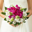 In this photograph, a romantic bouquet of magenta peonies and white alstroemeria is held as if ready for a wedding aisle in Clerkenwell, its colours alive in soft, natural daylight. The peonies sit at the centre like lush silk cushions, full and rounded, their many petals shifting from deep berry tones in the shadows to brighter magenta where the light touches. Encircling them, white alstroemeria unfold with a quiet grace, each bloom marked with delicate streaks of burgundy that feel hand-painted, giving the bouquet a refined, almost artistic character. Between these flowers rise slim spikes of pale green buds, not yet open, lending a sense of anticipation and renewal, like the early leaves you'd notice walking through nearby St James's Church Garden in spring. Generous greenery surrounds the composition, the leaves thick and slightly curved, offering both visual support and a cool, earthy backdrop that tempers the intensity of the magenta. The lighting is soft yet bright, catching the texture of each petal and the gentle sheen of the foliage, so that the bouquet appears almost three-dimensional, as if you could reach out and trace its contours. There is an imagined scent: faintly sweet from the peonies, fresh and leafy from the greens, suggesting a garden just after rain. The overall feeling is intimate and hopeful, ideal for a modern bride marrying in a Clerkenwell townhouse, or for someone sending flowers to celebrate a new chapter. Every detail speaks of careful craftsmanship and an understanding of how colour, texture, and form can quietly express love.