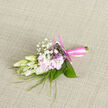 Laid gently on a soft, woven surface, this boutonnière appears like a thoughtfully composed miniature bouquet, crafted with the attentiveness you might expect from a florist in the quiet backstreets near St John's Square in Clerkenwell. At the centre, creamy ivory buds sit in that perfect moment before they fully open, their petals smooth and slightly curved, catching the soft ambient light. These are paired with blush-pink blossoms that open a little wider, their petals almost translucent at the edges, creating a romantic interplay between cream and pink. Fine blades of fresh green grass thread upward through the arrangement, adding a sense of movement and an almost wild garden touch, as though gathered from a hidden courtyard off Cowcross Street. Scattered throughout, tiny sprays of white baby's breath hover like lace, softening the outlines and lending a cloudlike lightness. The foliage behind the flowers is tidy and structured-short, broad leaves cradling the blooms and offering depth and contrast to the pastel colours. The stems are neatly bound together in a pale rose-pink satin ribbon, wound smoothly and secured with a precise knot, giving the design a polished, ceremonial feel. Light falls evenly across the piece, highlighting the satin sheen of the ribbon and the velvety texture of the petals, while casting gentle shadows that give depth. You can almost imagine a subtle, sweet fragrance rising from the flowers, the kind that lingers quietly as the wearer moves through a wedding ceremony or evening celebration. The overall impression is one of understated elegance and affection, a refined finishing touch for a groom or guest at a Clerkenwell celebration.