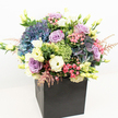 A luxurious, garden-style bouquet sits proudly in a sleek, matte black square box, photographed against a crisp white background so every colour sings. The arrangement is rounded and abundant, overflowing with large, cloud-like heads of hydrangea in deep periwinkle, dusty blue, and soft mauve, their petals densely packed to create a velvety, almost billowing landscape. Nestled between the hydrangeas are elegant lavender and blush pink roses, their spiral petals gently unfurling, adding a romantic softness and a hint of classic rose fragrance you can almost imagine. Creamy white lisianthus and buttery eustoma blooms weave through the design, some fully open with ruffled, trumpet-shaped flowers, others still in graceful buds that add height and a dewy freshness. Bright lime-green mossy clusters, reminiscent of Green Trick dianthus, bring playful texture and a contemporary edge, while delicate sprays of feathery pink astilbe float above the main blooms, like soft wisps caught in a light Clerkenwell breeze. Scattered sprigs of tiny fuchsia-pink fillers and subtle dark red berry-like buds create small points of contrast, supported by layers of lush, varied foliage in bright and deep greens that make the colours glow even more vividly. The minimalist black box grounds the palette, giving the design a modern London feel that would sit beautifully in a Clerkenwell loft, a design studio off St John Street, or an elegant townhouse near Exmouth Market. The overall mood is sophisticated yet joyful, perfect for marking a special birthday, a heartfelt thank you, or a quiet moment of support, the kind of premium arrangement often chosen for same-day flower delivery across EC1 when words need the gentle help of flowers.