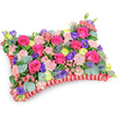 Seen from a gently elevated, oblique angle against a pure white backdrop, this pillow-shaped floral tribute reads as a comforting tapestry of colour and texture. The surface is densely filled-no green foam shows-so the eye rests on a field of hot pink roses whose velvety petals sit either as tight buds or begin to unfurl in soft spirals. Interwoven among them are clusters of shell-pink carnations, their ruffled edges lending a tender, fabric-like softness that echoes the cushion form. Bright lemon-yellow ranunculus punctuate the composition with sunlit accents, while deep purple lisianthus adds a cool, bell-shaped contrast that draws the gaze inward. Cool, spiky ears of silver-blue eryngium (thistle) push through the softer blooms, introducing an architectural, wild note that balances the arrangement's plushness. Rounded, silvery eucalyptus leaves and darker green fillers weave through the flowers, their matte sheen and faint menthol scent calming the overall palette. The whole design sits within a pleated, deep-red satin ribbon border that frames the rectangle like a ceremonial pillow, the ruffle catching soft highlights and grounding the colours. The light is even and gentle, bringing out petal texture and satin sheen without harsh shadow, as if arranged by hand in a quiet Clerkenwell workshop near Exmouth Market. This composition reads as a celebration of a life lived-joyful, tactile, and reverent-offering solace and a visual embrace at a time of remembrance without heavy mourning.