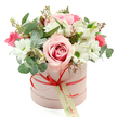 A sophisticated hatbox arrangement sits squarely in the centre of a white background, its soft colours and clean lines echoing the calm of a quiet Clerkenwell morning. The container is a smooth, cylindrical box in a muted pastel pink, finished with a coral-red satin ribbon wrapped neatly around its lower edge. At the front, the ribbon is tied into a tidy bow, from which hangs a rectangular cream gift tag printed in a gentle green script, suggesting a note of affection or a discrete florist signature. Above the rim, the flowers rise in a generous, domed bouquet that looks carefully measured but not rigid. The eye is first drawn to a striking pale pink rose with a deeper pink centre, fully opened to reveal its spiralling petals. Around it gather crisp white daisy-style chrysanthemums, each one centred with a bright green disc that punctuates the arrangement with a fresh, lively touch. Slender white alstroemeria, recognisable by their trumpet shape and subtle speckling, thread between the larger blooms, while small, vivid deep pink flowers-resembling spray roses or lisianthus-add pockets of vibrant colour that keep the design feeling joyful rather than overly soft. Eucalyptus foliage with round, silver-green leaves arcs gently outward, catching the light with its matte, slightly bluish tone, while darker green filler leaves create a textured backdrop. Dainty stems tipped with tiny green buds and a few reddish-brown sprigs lend a slightly wild, garden-inspired character. It is the kind of elegant hatbox bouquet a local florist might prepare for delivery to a loft near Farringdon Station or a cosy flat off Rosebery Avenue, ideal for marking a birthday, anniversary, or heartfelt thank-you with understated luxury.