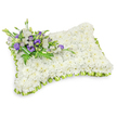 A tender rectangular sympathy pillow, entirely surfaced with hundreds of small, pristine white chrysanthemum heads, lies against a clean white backdrop with a soft drop shadow that defines its gentle, puffy profile. The chrysanthemums are packed closely to form a cloud-like, velvety carpet of blossoms whose subtle tea-like scent and powdery petal texture suggest quiet rest and consolation. Around the pillow's edge a soft, lime-green satin ribbon has been pleated into a continuous scalloped trim, the cool green offering a fresh, hopeful outline that lifts the whiteness without intruding on its serenity. In the upper left corner a composed spray of accent flowers anchors the design: crisp white lisianthus with layered, cup-like petals nestle beside deep purple lisianthus buds and open blooms, their mauve-lilac tones providing a respectful, dignified contrast. Sprigs of feathery fern fronds and glossy dark green leaves weave through the cluster, while a broader leaf wrap at the base holds the stems in an understated, natural bind. The lighting is soft and even, emphasising the pillow's three-dimensional curve and the tiny shadows between chrysanthemum heads, so the arrangement reads as both tactile and composed. This design reads as a personal, comforting tribute-suitable for a bedside service, graveside laying, or a quiet memorial-expressing peace, reverence, and the warmth of remembered presence. Imagined for services in and around Clerkenwell, the piece feels like a delicate, hands-on remembrance crafted to soothe those who gather to say goodbye.