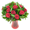 A lavish, rounded bouquet of roughly a dozen deep crimson roses fills the frame, each bloom opened into a perfect spiral of velvety petals whose colour reads almost wine-dark at the heart and brightens toward the petal edges. The roses sit closely clustered to form a dense, domed profile; soft, even studio light skims the petals, revealing fine veining and a subtle satin sheen that suggests freshly cut stems. Nestled between the heads is an abundance of dark green foliage - glossy, flat leaves like Ruscus or Salal form a rich, verdant backdrop while broader, emerald Aspidistra-style leaves are expertly curled and rolled into sculptural loops and tubes. Those folded leaves create rhythmic arcs that cradle each rose and add a contemporary architectural counterpoint to the classic blooms.  The arrangement is anchored in a tall, slender translucent ruby-red glass vase with a faintly ribbed, twisted texture that catches the light in vertical highlights, echoing the roses' passionate tone. Set against a clean white background, the strong reds and deep greens pop with clarity; the composition reads as both intimate and formal, a floral statement that radiates romance and refinement. Imagine the faint, warm scent of rose and green leaves, and the way this piece would command a dining-table centre or an anniversary gift delivered across Clerkenwell. The bouquet's balance of traditional red roses and modern foliage styling makes it equally suited to Valentine's Day, an elegant proposal, or a quiet, heartfelt celebration in a nearby courtyard off Exmouth Market.
