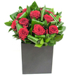 In this elegantly modern arrangement, rich velvety crimson roses form the unmistakable heart of the display, each bloom opening in lush, generous spirals that read like small, satin sculptures. The roses are carefully clustered so their deep red faces press close together, while broad glossy green leaves are looped and curled around them to create a rhythmic frame-each leaf folded with intent to add movement, shine, and a sculptural silhouette. The composition sits low in a matte black cube that gives the bouquet a confident, geometric base; its smooth, flat surface contrasts with the soft petals and the slightly waxy sheen of the foliage, grounding the design with contemporary poise.

Bright, clean lighting picks out highlights on the petals and catches the gleam of water on the inner leaves, lending an almost tactile clarity as if you could run a fingertip along the petals and feel their silken softness. Close to the arrangement there is an airy, fresh green scent layered beneath the classic heady perfume of rose-a subtle, uplifting note that balances the bouquet's romantic intensity. As a florist familiar with Clerkenwell's quiet courtyards and the lively stalls of Exmouth Market, I imagine this piece sitting on a reception desk in a nearby design studio or as the centrepiece for an intimate anniversary dinner on St John's Square.

Crafted with intention, this bouquet reads as both an urban statement and an intimate gesture-perfect for romantic milestones, sincere thanks, or any moment that calls for passionate, refined floristry made for modern London interiors.