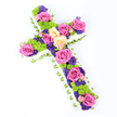 On a pure white background, a striking floral cross lies at a slight diagonal, its traditional shape softened and enriched by layers of carefully arranged flowers. The outer edge of the cross is formed by a dense border of crisp white spray chrysanthemums, each tiny bloom sitting snugly next to the next to create a cloud-like frame that gently blurs the hard lines of the shape. Just beneath this, a slim ribbon of light green-either folded satin or tightly pleated foliage-curls neatly around the perimeter, revealing itself in small flashes of fresh, springlike colour and giving the tribute a polished, hand-finished look. Inside this border, the cross is filled to overflowing with colour. Large roses in shades of bold hot pink and creamy pale yellow dominate the design, their luxurious, full-petalled heads placed to draw the eye along the vertical and horizontal beams. Between them, rounded lime-green button chrysanthemums are tucked in clusters, bringing a lively texture and a contemporary freshness that contrasts beautifully with the classic roses. Interspersed among these larger flowers are smaller blossoms in deep purple and violet, likely lisianthus or statice, which introduce notes of shadow and depth, like quiet corners in a Clerkenwell side street at dusk. The entire arrangement is evenly lit, with no harsh shadows, so every petal and ribbon fold is clearly visible against the white ground. This is a thoughtful funeral or memorial cross, the sort of piece chosen for services across Clerkenwell and nearby neighbourhoods, expressing condolence, Christian faith, and a gentle sense of peace through its balanced palette of pink, cream, green, and purple blooms.
