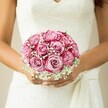 The moment feels suspended in time-a young bride, framed by the gentle light streaming through Clerkenwell streets, delicately embraces a bouquet that breathes timeless romance. Lush pink roses dominate, each one swirling from the palest blush in the centre to vibrantly deep magenta at the petal's edge. The gradient in the blooms echoes the painterly tones of Clerkenwell's local galleries, creating visual harmony and intrigue. Frothy clusters of snowy gypsophila weave between the roses, their softness reminiscent of the first spring morning mist hovering above Farringdon's rooftops. Her hands, gracefully tanned and adorned with subtle, light-shimmering nails, cradle the bouquet just over the flowing lace-emroidered bodice of a white gown, the V neckline adding to the dress's elegant silhouette. The loosely draped sash to the right introduces movement and a hint of whimsy. In the blurred neutral backdrop, the bouquet commands every bit of attention, emanating a joyous anticipation and hope. It is not merely an arrangement-it is an embodied promise from Clerkenwell Flowers to craft moments that linger in memory, fitting for couples whose love story is as unique as the neighbourhood itself.