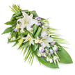 An elongated memorial spray of white and green, accented with soft lavender, stretches diagonally across a plain white background, its form reminiscent of a tender farewell laid carefully by hand. The upper section is lush with open oriental lilies, their wide, ivory-white petals unfurling like calm waves, some revealing pale stamens at the centre, others still partially closed, holding their fragrance in reserve. Nestled among the lilies are clusters of lisianthus, their smaller, layered blooms offering a gentle contrast of texture. Many of these flowers are pure white, while others show delicate lavender or light purple tracing along the petal edges, adding a hushed note of colour that calls to mind early evening shadows around Clerkenwell Green or the quiet paths of nearby Spa Fields. Countless lisianthus buds, round and tightly wrapped, dot the arrangement and lend it depth. A robust foundation of foliage anchors the spray: broad, dark green leaves provide weight and substance, lying almost like a protective cloak beneath the blossoms. Some leaves have been artfully rolled and folded into contemporary, ribbon-like shapes that step the eye slowly down the design. From the right, long, slender palm fronds extend out and away, creating a sense of movement and soft release, as if the flowers were gently guiding the gaze onwards. The interplay of smooth petals, crisp foliage, and layered greens suggests both structure and tenderness. This is the kind of carefully balanced tribute a local Clerkenwell florist might prepare for a funeral or cremation service, suitable for display in a chapel, at a graveside, or on a casket to express sympathy, remembrance, and enduring love.