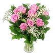A clear glass vase stands at the centre of a white, uncluttered background, holding a full, rounded bouquet of pink roses and white baby's breath that immediately evokes softness and welcome. Around ten to twelve roses form the main structure, their petals in shades of gentle blush to richer pink, arranged so that no bloom dominates, but rather all work together in a harmonious circle. Some roses are completely open, showing intricate swirls at the core, while others are only partially unfurled, lending the piece a sense of freshness and promise that feels fitting for baby congratulations and early milestones. Between the larger flowers, airy streams of Gypsophila weave through, each spray carrying numerous miniature white blossoms that appear like floating specks of light against the deeper tones of the petals. Dark green foliage sits beneath and around the roses, its leaves smooth, layered, and slightly glossy, giving a supportive frame that grounds the more delicate elements. Through the transparent sides of the vase you can see the straight green stems, neatly gathered in clear water, a small but telling detail of the bouquet's just-prepared nature. Soft, balanced lighting from above and slightly to the side gives the petals a velvety look, highlighting their gentle curves and subtle colour shifts while keeping the overall mood calm and serene. The composition feels timeless yet contemporary, easily at home on a mantlepiece in a Clerkenwell apartment overlooking St John's Square, on a reception desk in a local studio, or delivered as a thoughtful gift to new parents welcoming their baby in one of the nearby hospitals or clinics.