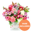 A romantic mixed bouquet sits proudly in the centre of a white backdrop, its colours and textures carefully composed to evoke affection and celebration, as if created for a surprise delivery to a flat or studio off Rosebery Avenue in Clerkenwell. The flowers are arranged in a clear, square glass vase, whose base is encircled by a decorative white band printed with small, bright red hearts and the handwritten phrase all my love, giving a distinctly intimate message even before the card is read. From the vase, a full, rounded arrangement rises, dominated by pink roses in several shades: some petals glow with deep fuchsia and magenta tones, others soften into a gentle pastel pink. Their velvety blooms appear layered and lush, some fully open and others still partially closed, suggesting the bouquet will continue to unfold over several days. Interwoven with the roses are generous carnations in coral and peach-pink hues, their finely ruffled petals creating a soft, cloud-like texture. Threaded throughout are small daisy-style chrysanthemums in lavender and purple, each with a bright lime-green centre that adds a lively spark of freshness. Creamy white carnations and petite white daisy-like blossoms break up the richer colours, ensuring the overall effect remains light and uplifting rather than heavy. Around the blooms, an abundance of foliage adds structure and depth: rounded eucalyptus leaves in silvery-green tones, darker green leaves providing contrast, and a few reddish-brown sprigs giving a subtle hint of seasonal warmth. The neatly cut stems are visible through the vase, showing the care taken in the design. In the lower right corner of the image, a circular orange label with bold white capitals announces VASE INCLUDED, clearly indicating that this bouquet arrives as a complete, ready-to-display gift for any special moment in Clerkenwell.