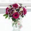 A vivid mixed bouquet of fuchsia gerbera daisies, creamy white roses, and purple-tipped chrysanthemums stands proudly in a short, clear glass vase against a softly glowing white background, as if placed on a bright Clerkenwell windowsill. The large gerberas, in deep magenta with dark central disks, draw the eye immediately, their broad petals radiating out like little suns. Around them, clusters of daisy-like chrysanthemums show off petals dipped in rich purple that fade toward clean white edges, each flower circling a cheerful yellow centre. Creamy spray roses, some fully opened and others still in tight buds, nestle between the bolder blooms, bringing a gentle, romantic softness. Fine sprigs of white gypsophila thread through the arrangement, their tiny cloud-like blossoms giving a light, airy texture that contrasts with the velvety petals and glossy greenery. Deep green leaves and stems provide structure and a natural frame, their fresh colour suggesting the bouquet has just been prepared by a local florist. The glass vase itself is simple and jar-like, its textured surface catching the light, and a rustic bow of natural twine is tied neatly around the neck. From the twine hangs a small pale gift tag with a pink floral motif and flowing script, hinting at a personal message for a birthday, congratulations, or a just-because surprise delivered to a home or studio near Exmouth Market. Soft daylight filters from behind, illuminating every petal and leaf, so the bouquet seems to glow with quiet celebration, capturing the feeling of receiving fresh flowers in the heart of Clerkenwell.